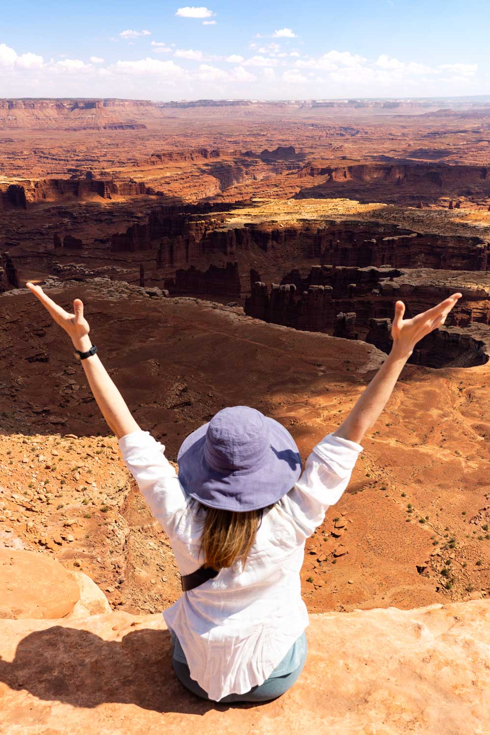 Female hiker in blue hat and hands up in the air, sitting on the edge of a large desert canyon with red rocks extending to the horizon