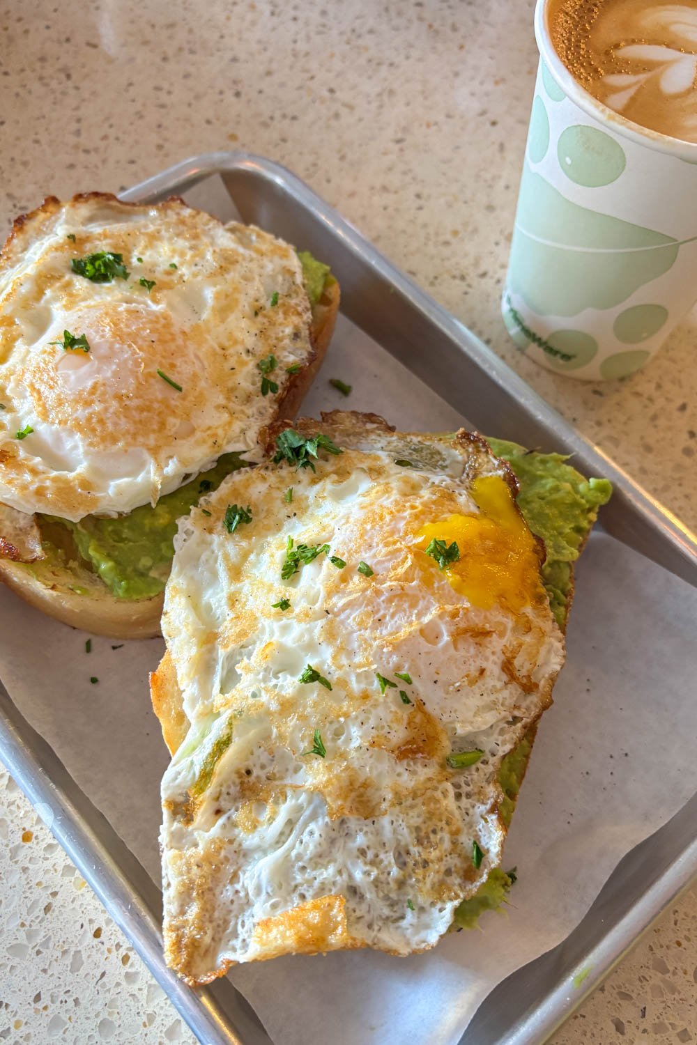 Metal tray-style plate of avocado toast with fried eggs and a takeaway coffee cup