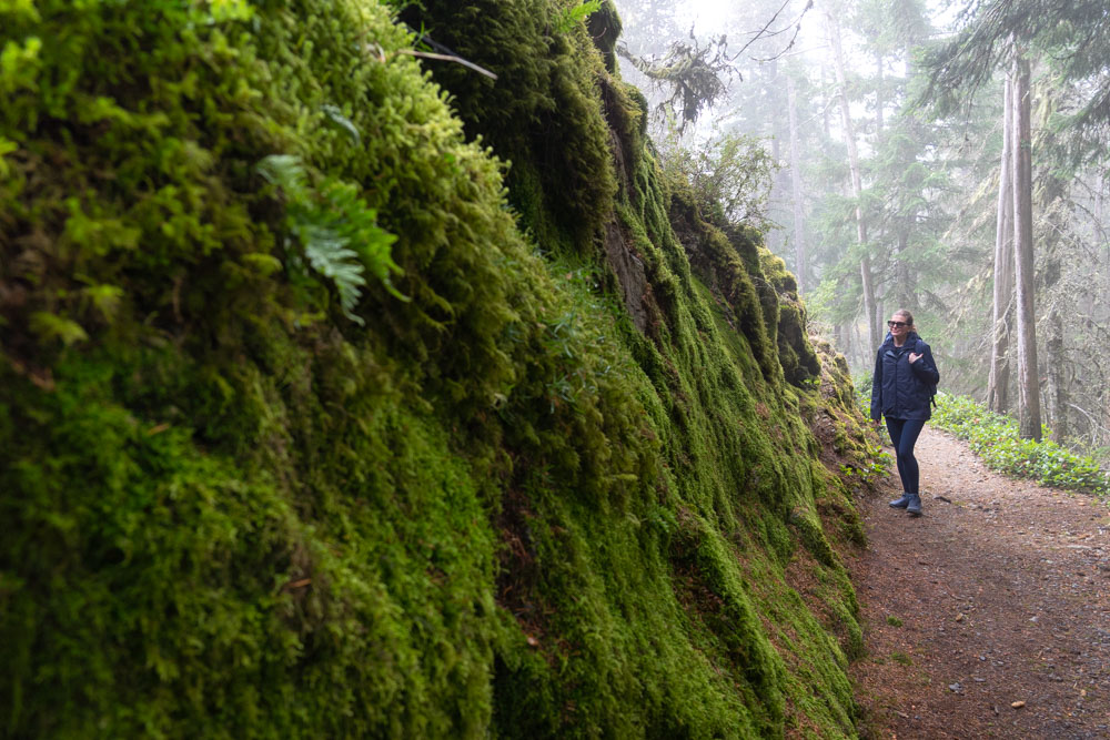 Woman standing next to large rocky wall covered in moss, in forest