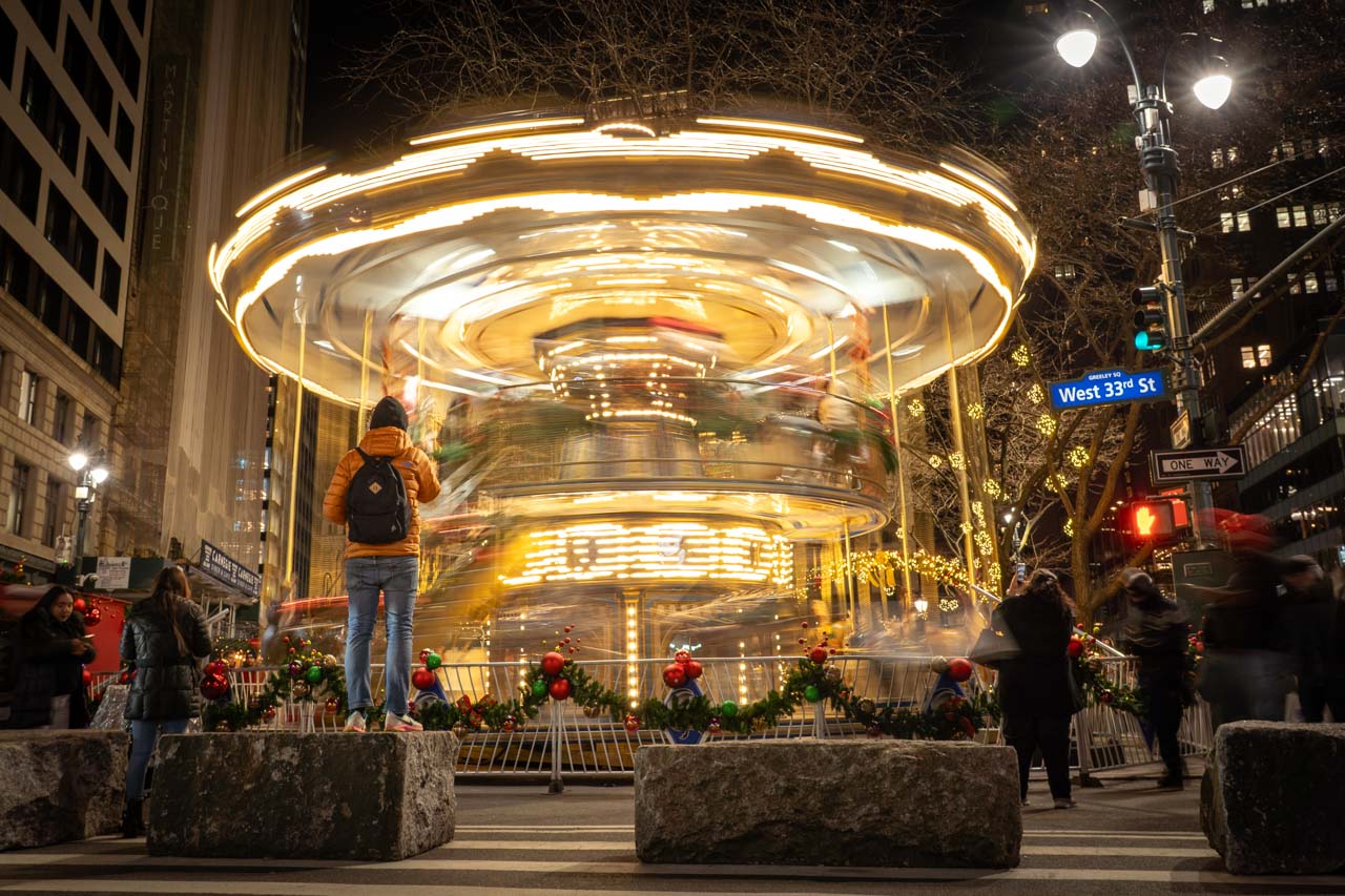 The blurred lights of a double-decker carousel turning at night on a New York City street, with onlookers taking photos