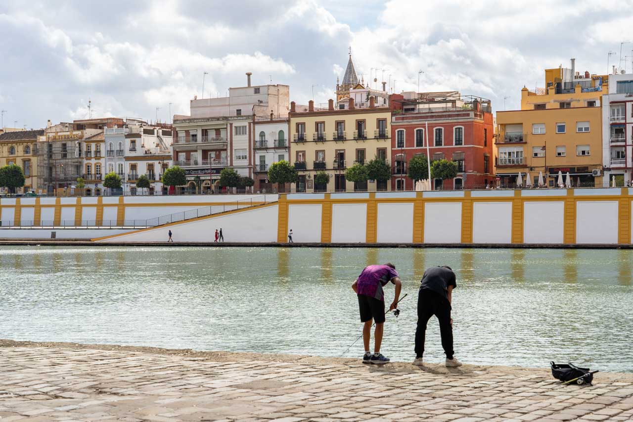 Two young boys with a fishing rod, looking down at the Guadalquivir River with Triana buildings in the background