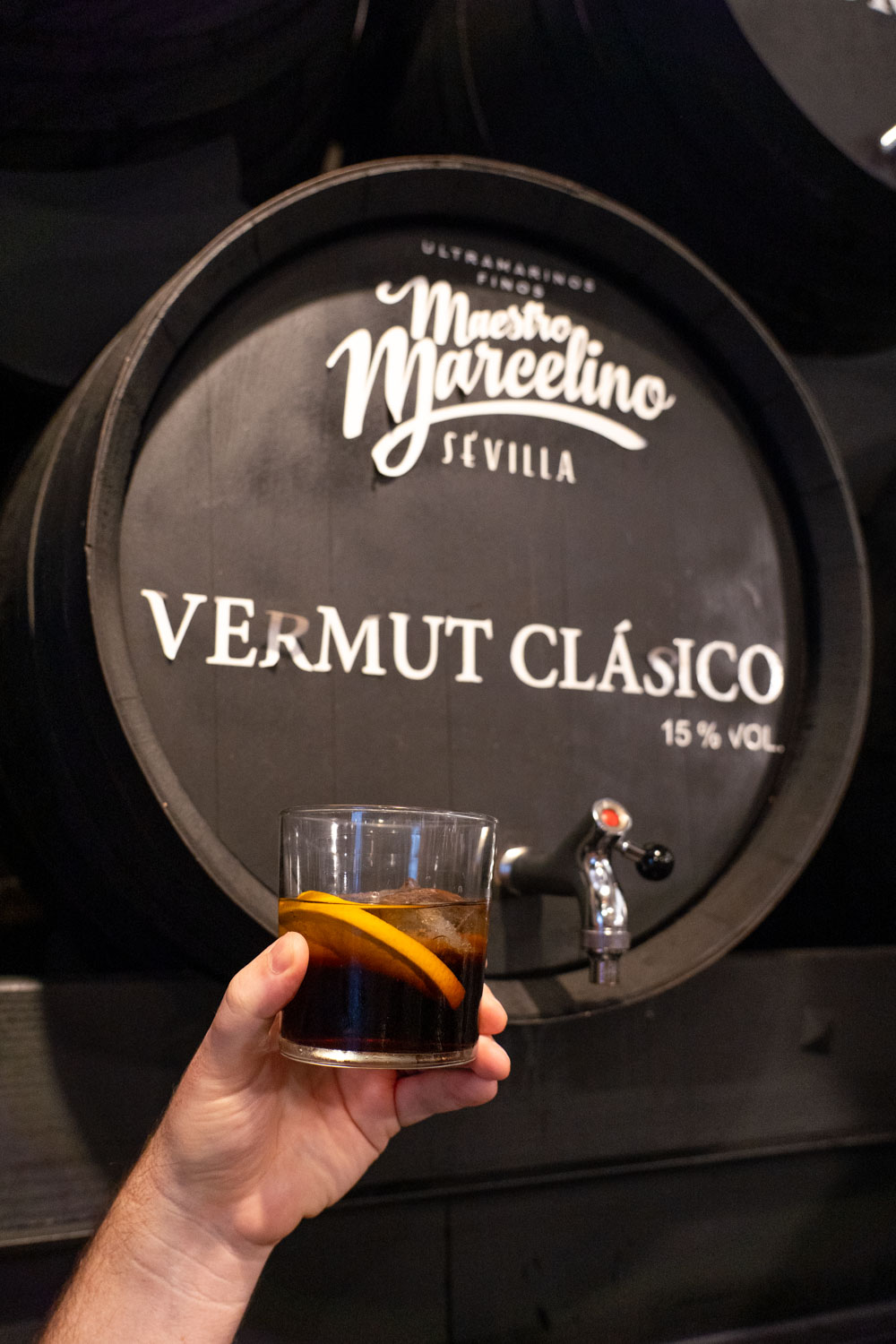 Glass of vermouth with orange slice being held infront of black barrel reading "Vermut Calsico"