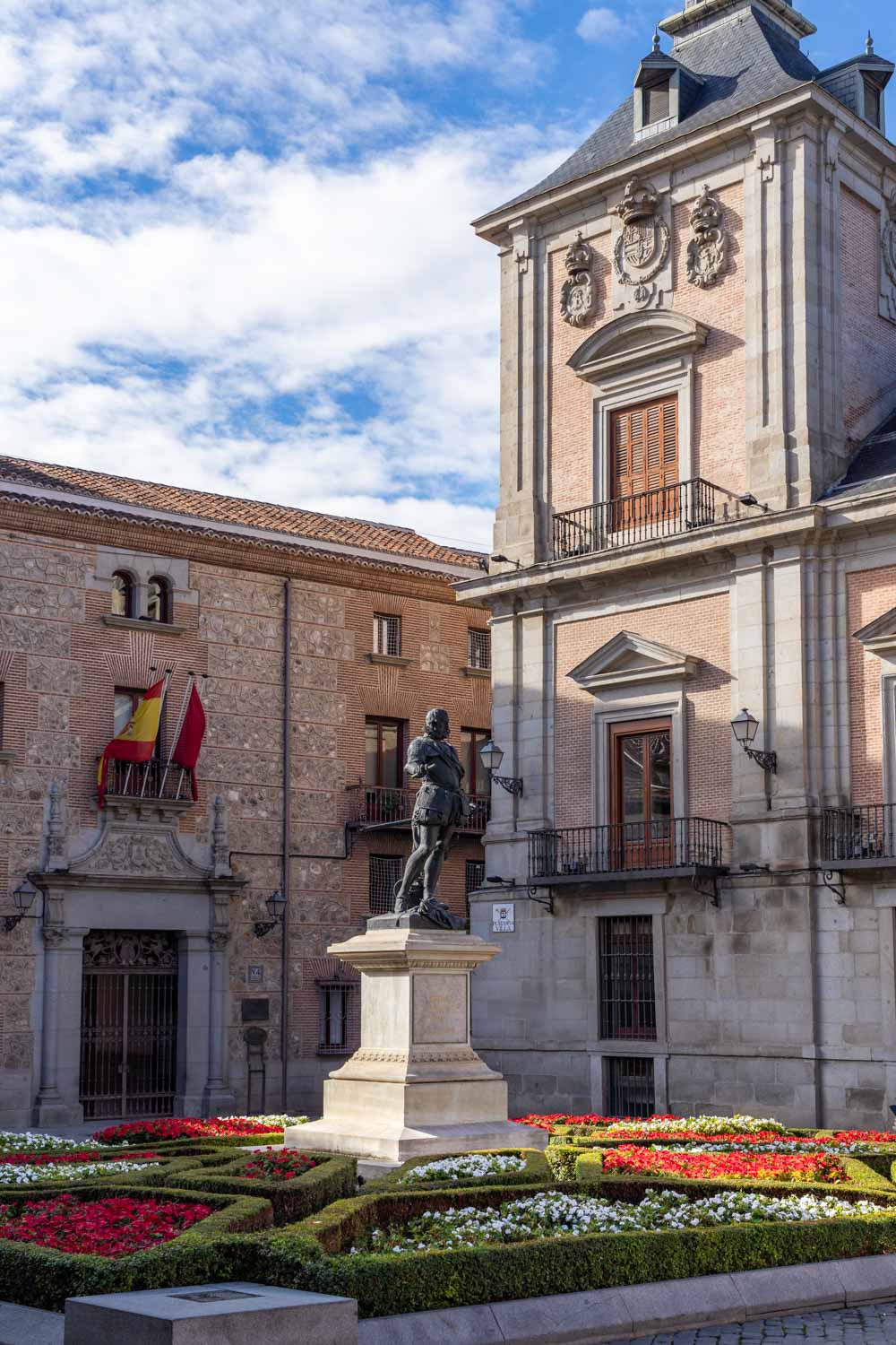 Image of Plaza de la Villa, in mid-morning sunlight, with blooming garden beds at the foot of a statue of Álvaro de Bazán
