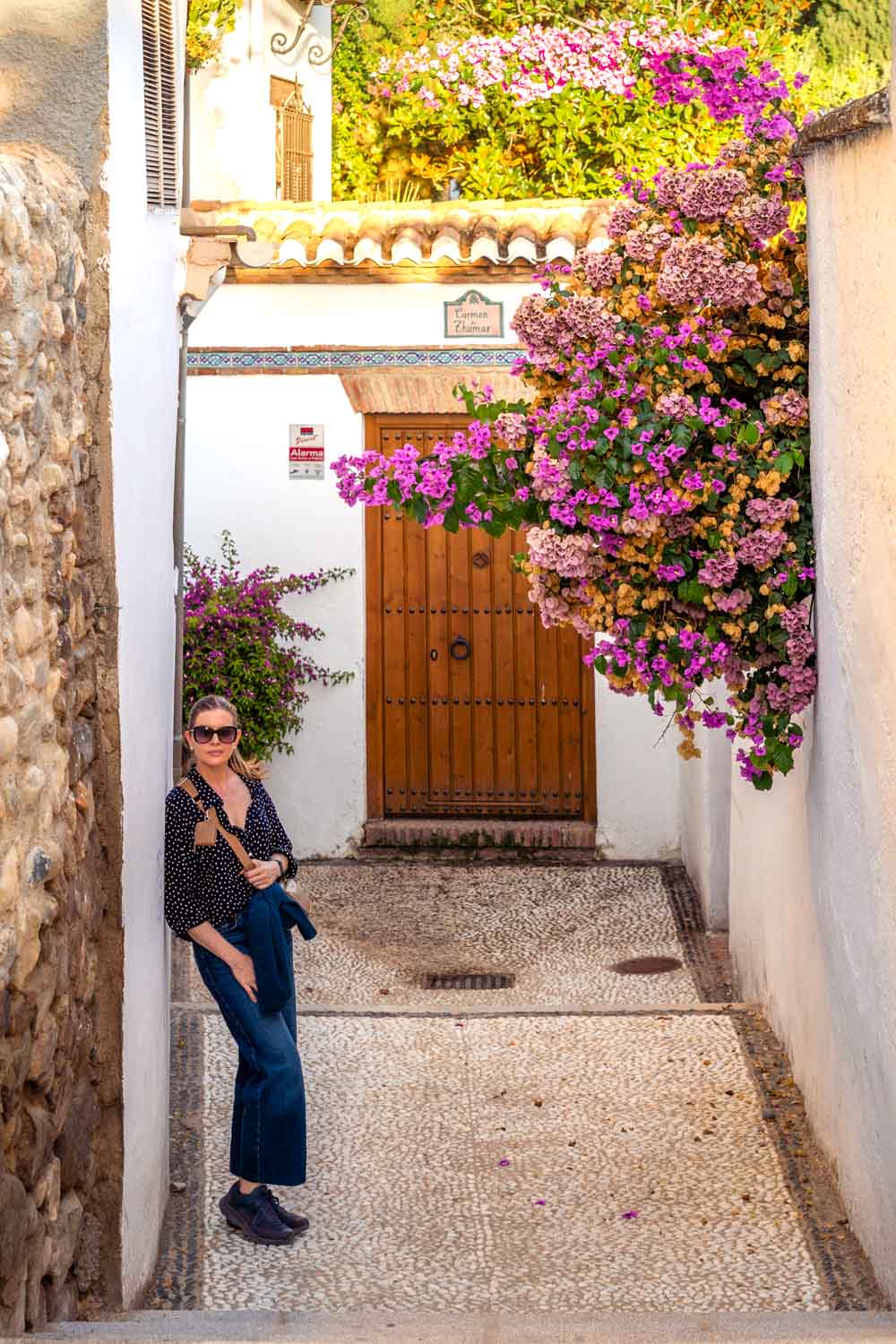 Woman leaning on the wall of a narrow street with a wooden door in the background and pink bouganvilleas growing over the wall