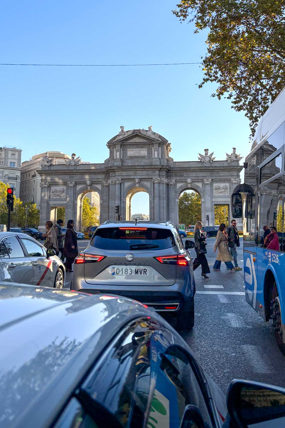 Pedestrians crossing at traffic light in Madrid, backed by ornate city gate and blue sky