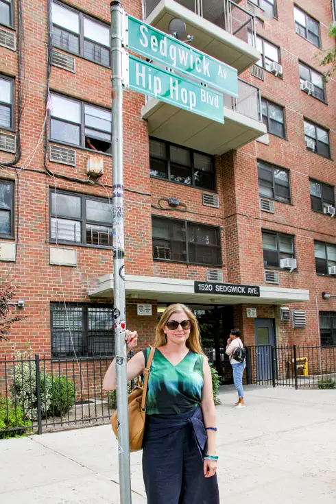 Woman leaning against street sign reading Sedgwick Av and Hip Hop Blvd