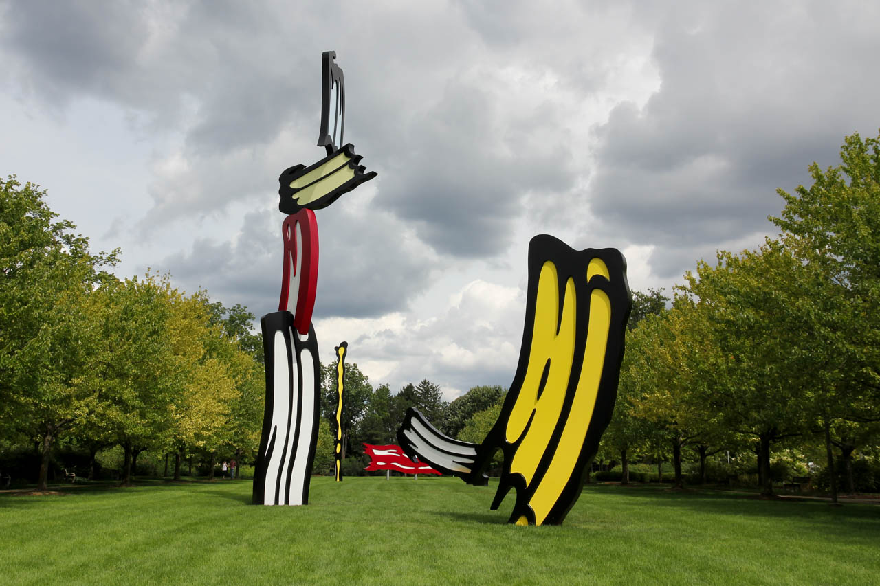 Giant brushstroke sculptures in yellow, white, red and black on lawn bordered by trees