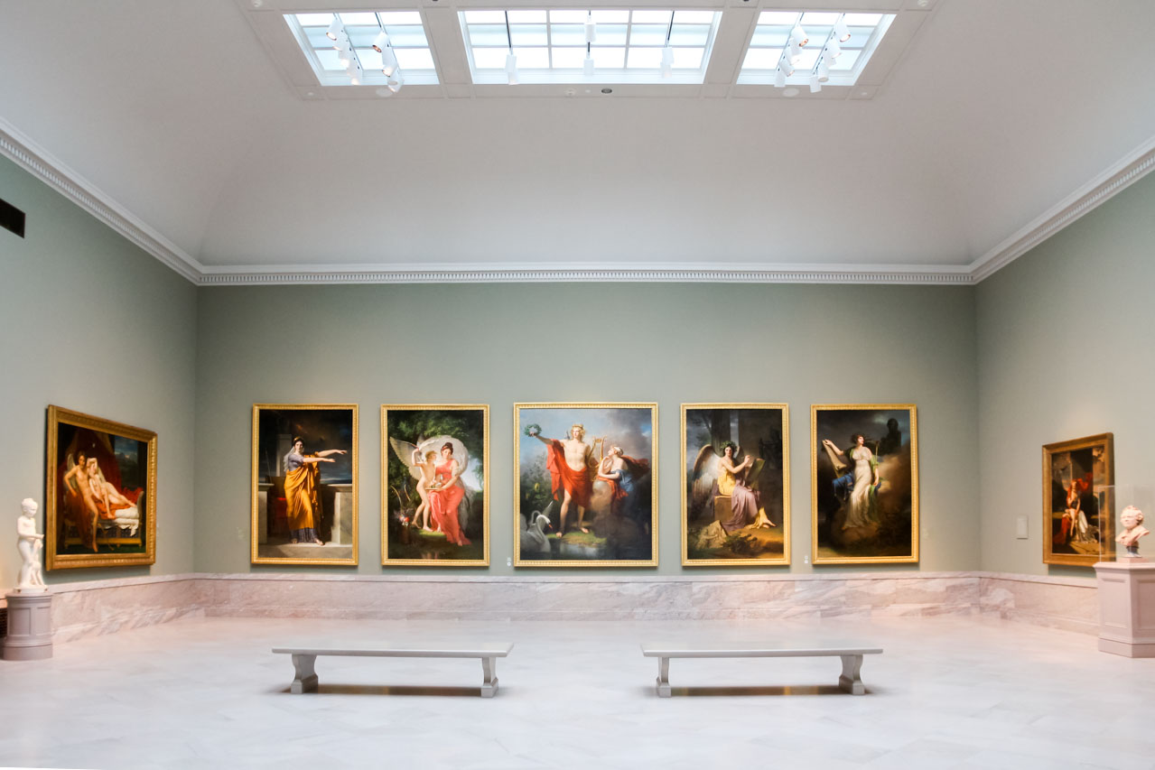 Series of paintings on a green wall with marble floor and benches in foreground