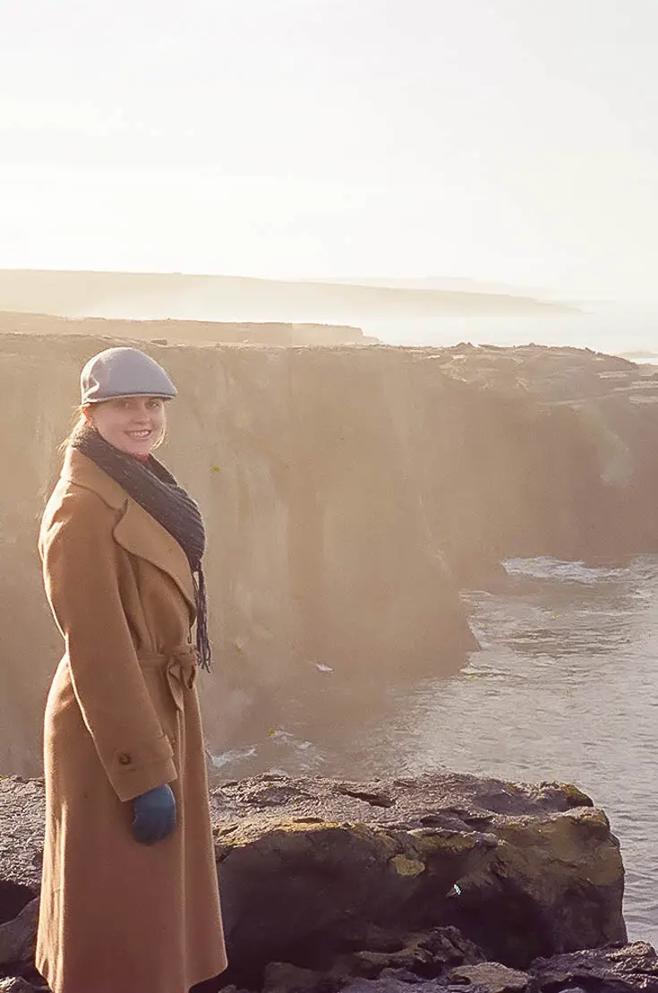 Woman in tan coat and grey hat standing on the edge of the Cliffs of Moher