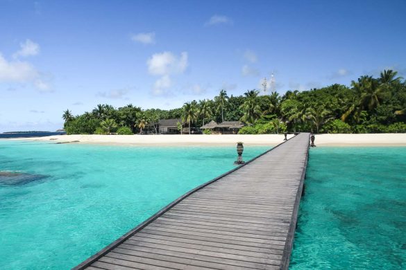 Photo of a wooden jetty leading to a sandy, tropical island surrounded by turquoise waters in the Maldives