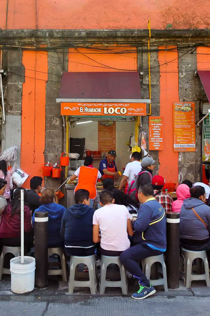 People seated on small plastic stools crowded around an eatery with bright orange walls and a service window onto the street