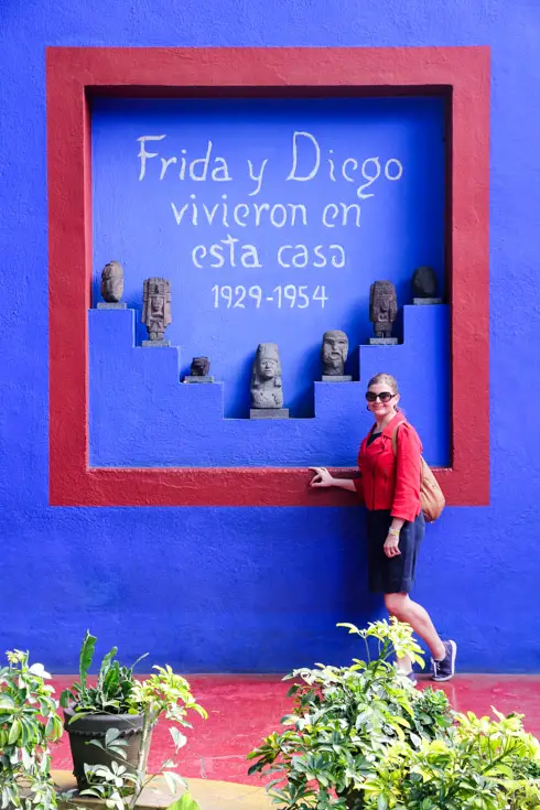 Woman standing infront of cobalt-coloured wall with niche holding sculptures and painted text reading "Frida y Diego vivieron en esta casa 1929-1954"