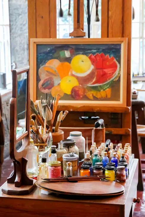 Frida Kahlo's studio with unfinished painting of fruit on an easel, and paints in foreground