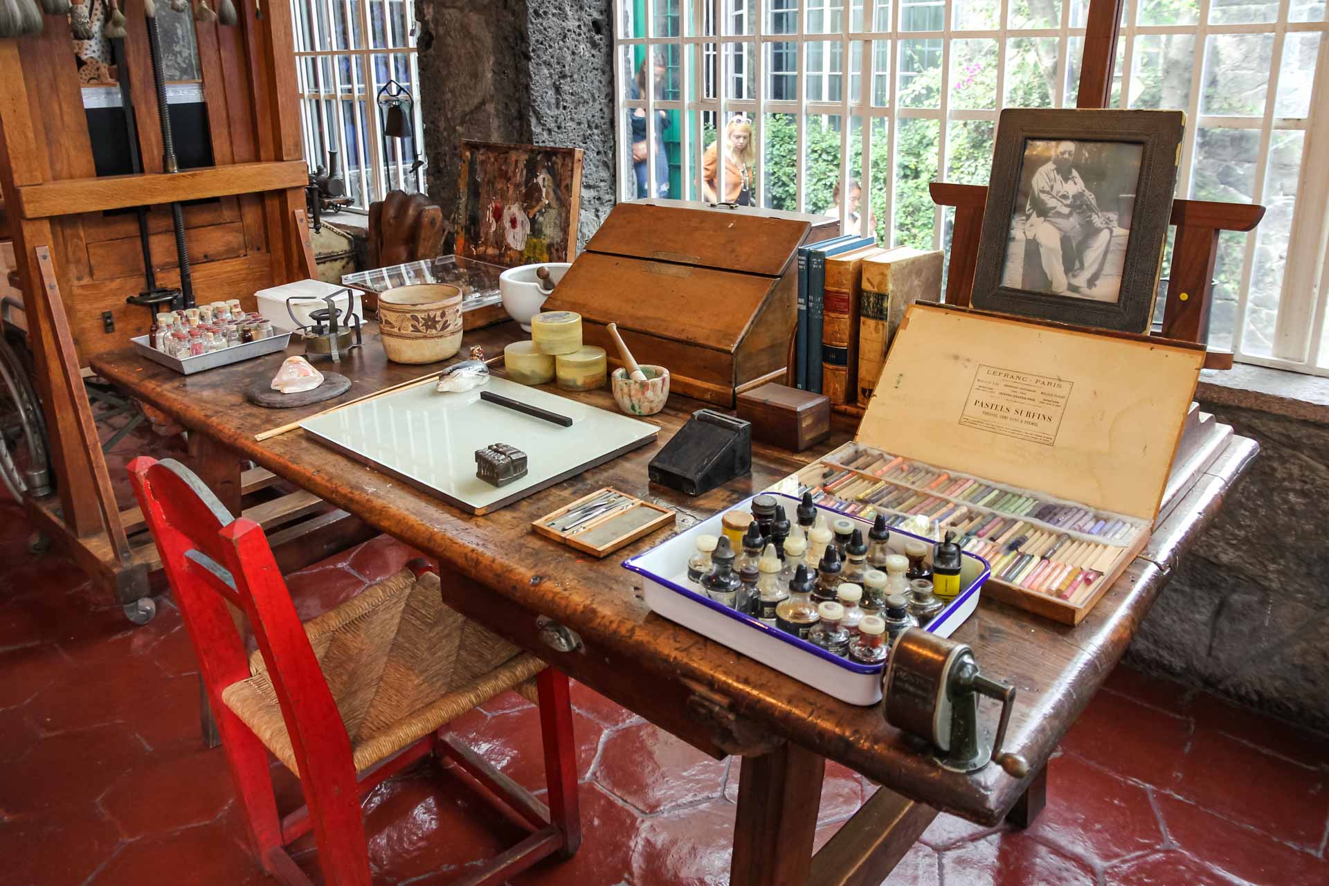 Frida Kahlo's studio at The Blue House with paints and pastels on a wooden desk, with a framed portrait of Diego Rivera