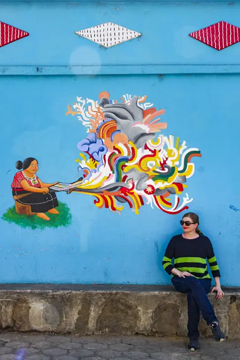 Woman sitting infront of blue wall with mural of Guatemalan women weaving on backstrap loom