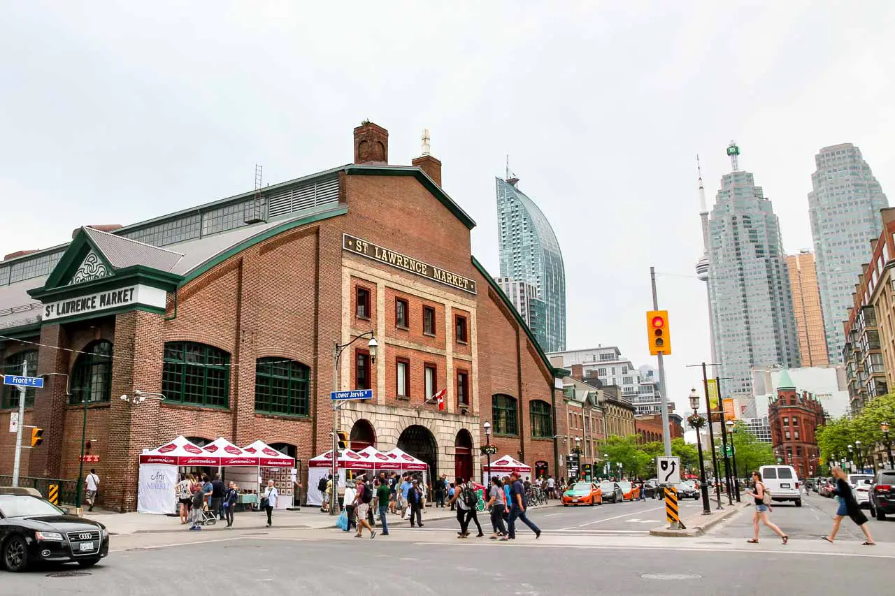 Exterior of large, St Lawerence Market building in red brick, viewed across a traffic intersection with city buildings including Gooderham flatiron in background