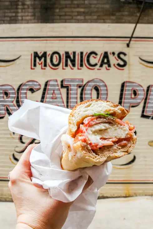 Hand holding sandwich in front of Monica's Mercato sign painted on side of brick building