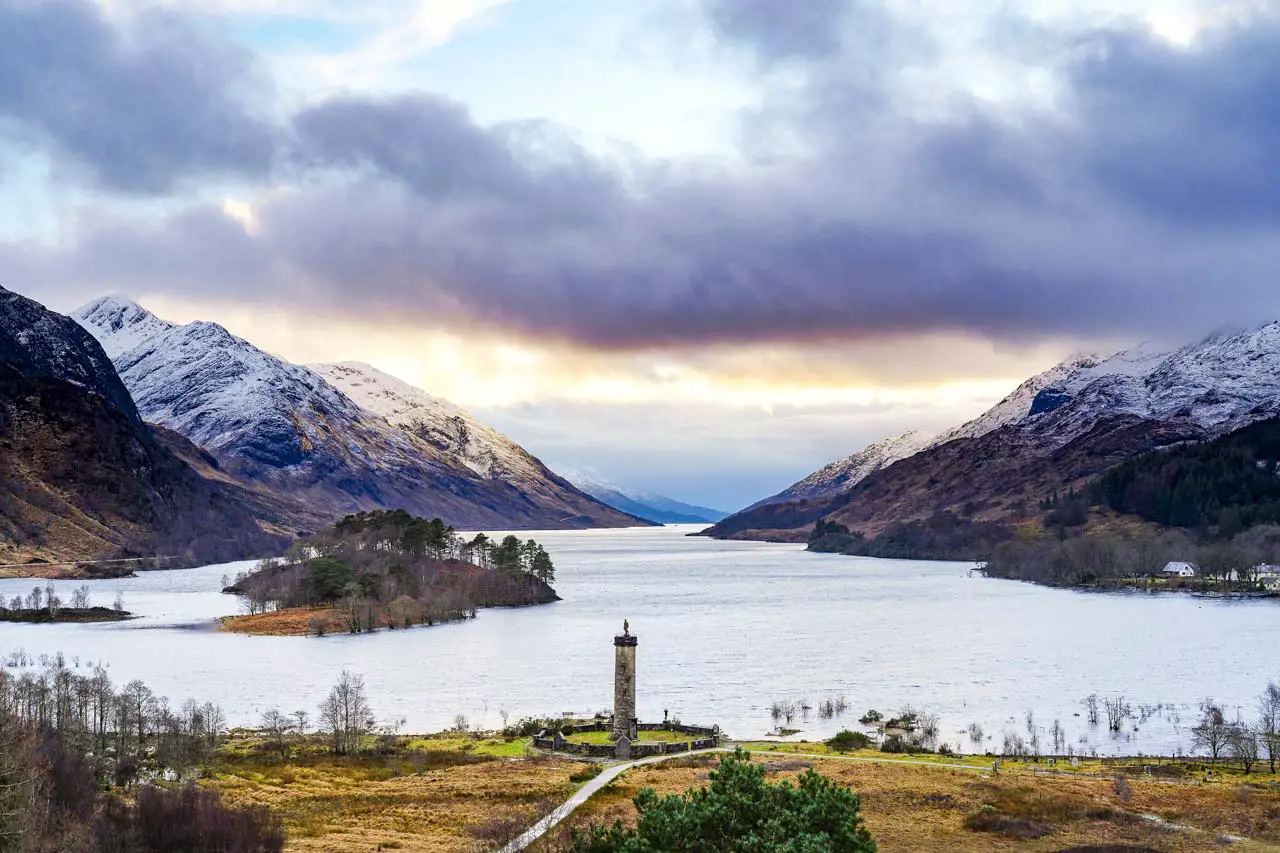 View looking down over Glenfinnan with monument in foreground and loch extending to the horizon. Mountains are illuminated by a colourful sunset.