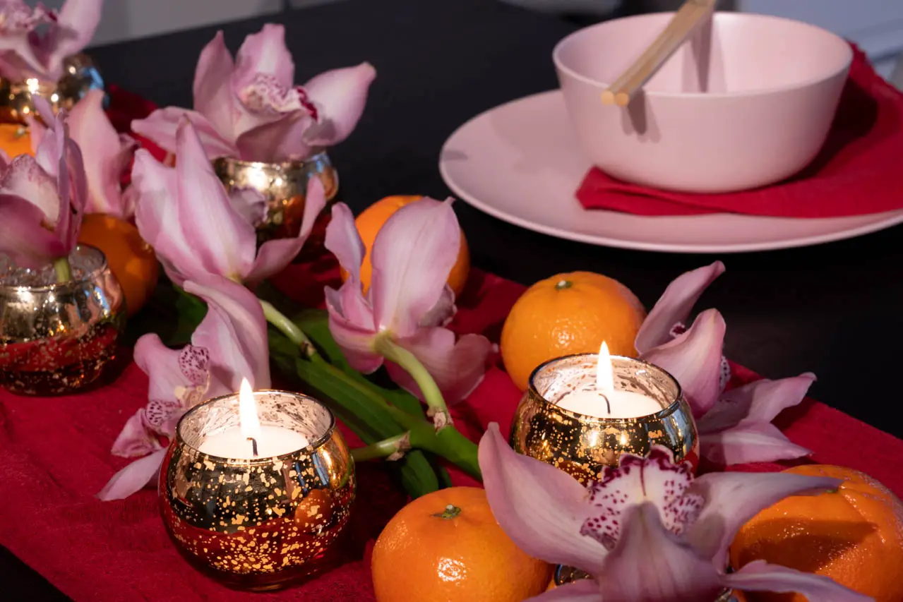 Mandarins, pink orchids and gold tealight candles on a red table runner, with a pink bowl and chopsticks in the background.