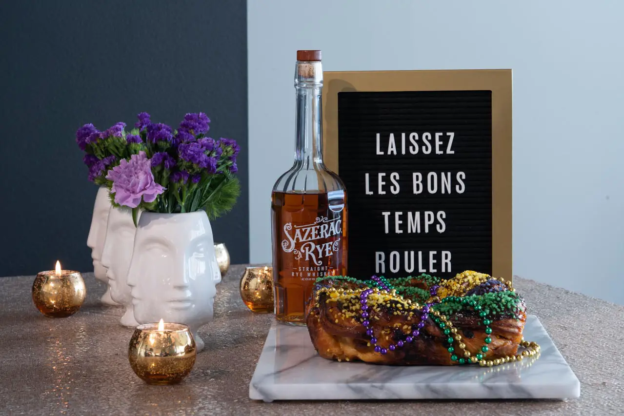 Sign reading "Laissez les bons temps rouler" with a bottle of Sazerac Rye whiskey, king cake on a marble platter and purple flowers in white vases with faces.