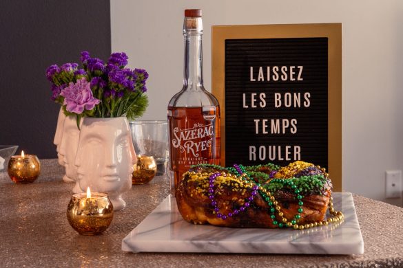 Sign reading "Laissez les bons temps rouler" with a bottle of Sazerac Rye whiskey, king cake on a marble platter and purple flowers in white vases with faces.