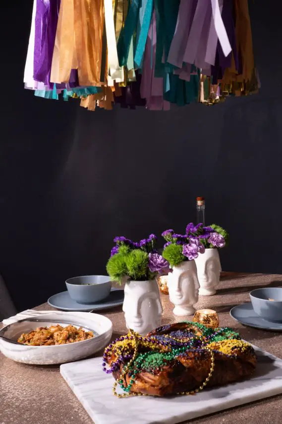 King Cake and bowl of Jambalaya on a table with gold sequin tablecloth and face vases of purple and green flowers.