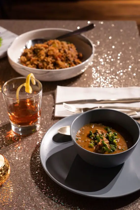 Bowls of gumbo and jambalaya with a cocktail on a gold sequin tablecoth.