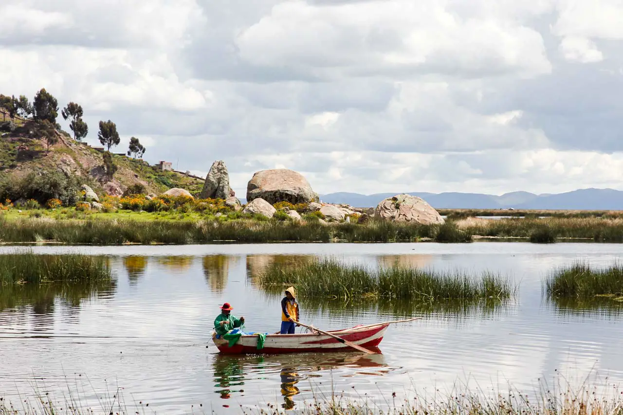Two people in a rowing boat among reeds on a lake with boulders in the background