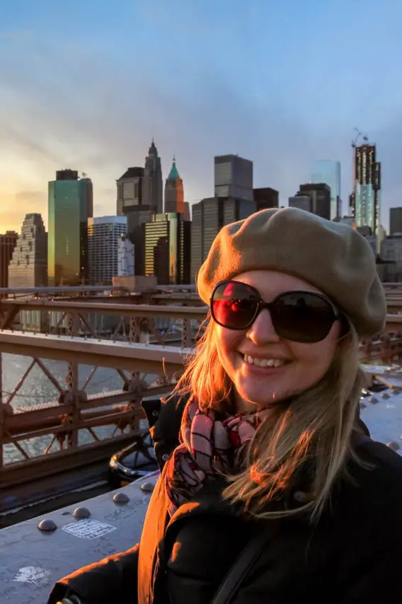 Madam ZoZo on Brooklyn Bridge, wearing beret, standingon Brooklyn Bridge at sunset with Manhattan skyline in background.