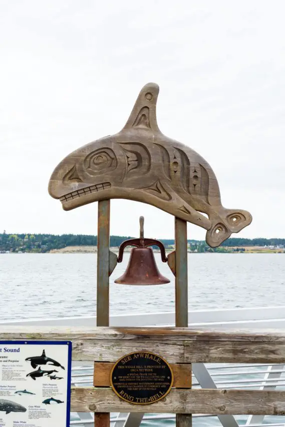 Indigenous wood carving in the shape of a whale with a lare bell hanging below, facing the water
