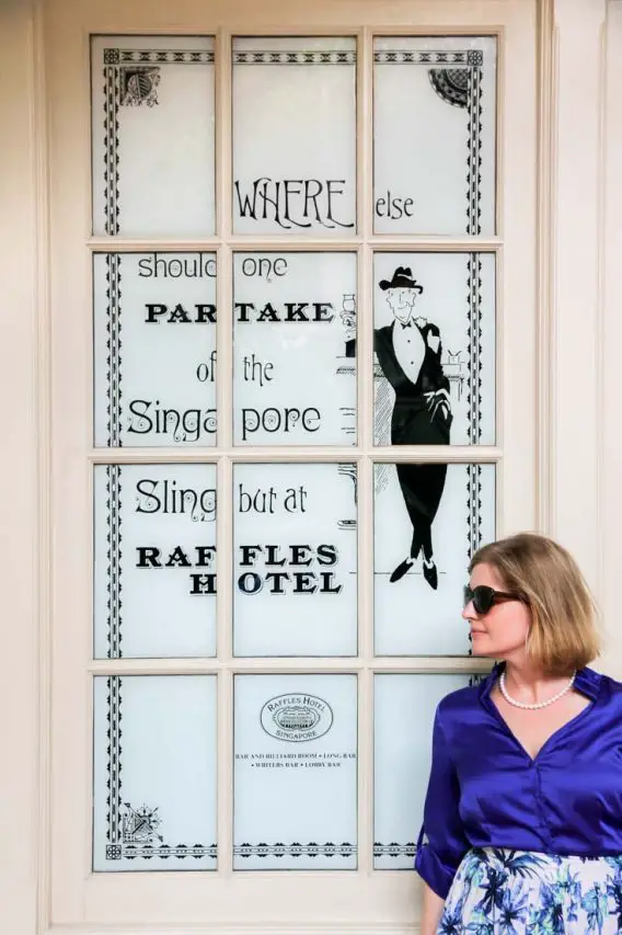 Madam ZoZo standing in front of a window sign that says "Where else should one partake of the Singapore Sling but at Raffles Hotel"