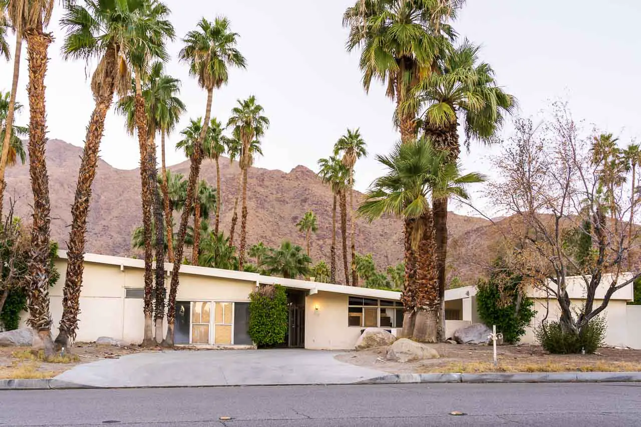 Run down Mid-Century Modern home in cream, with palm trees and mountains in the background