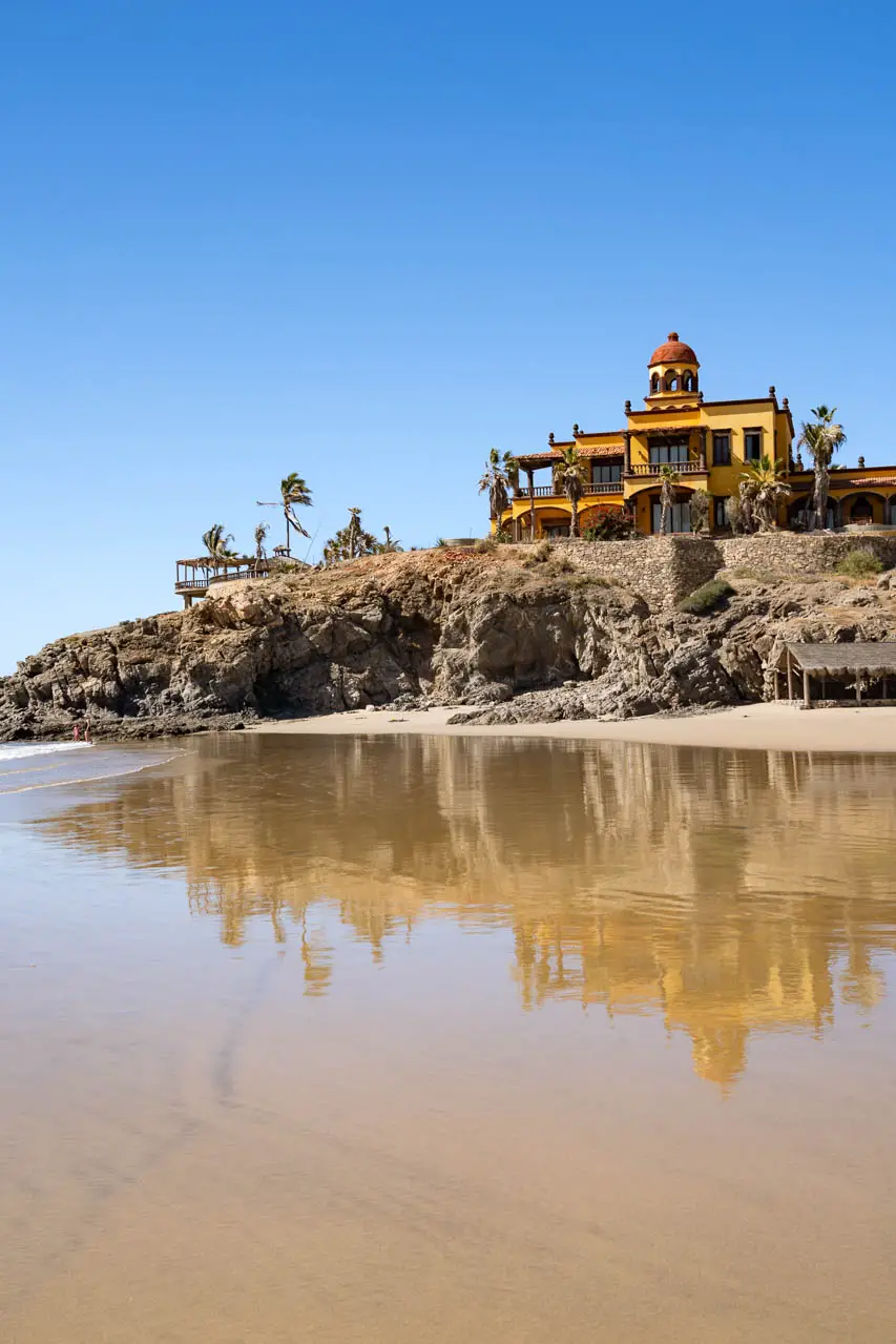 Large yellow Spanish colonial building atop a rocky cape, reflected in wet sand on the beach