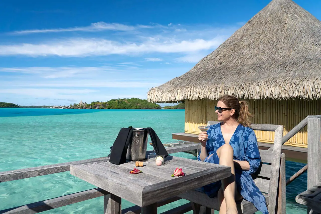Woman in blue dress, holding a champagne glass and looking out at turquoise, Bora Bora lagoon.