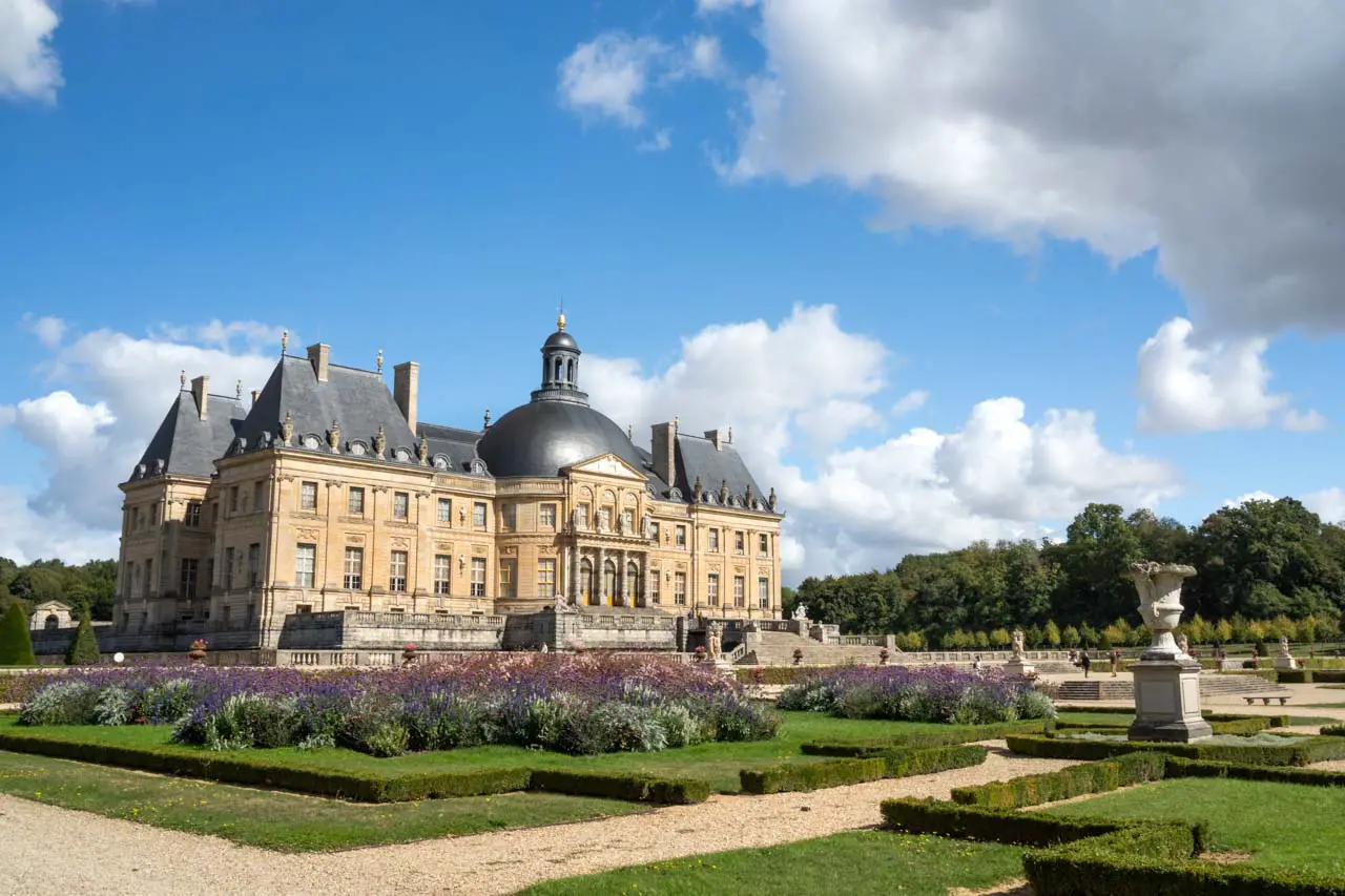 Image of a French Baroque castle exterior and formal gardens on a sunny, blue sky day