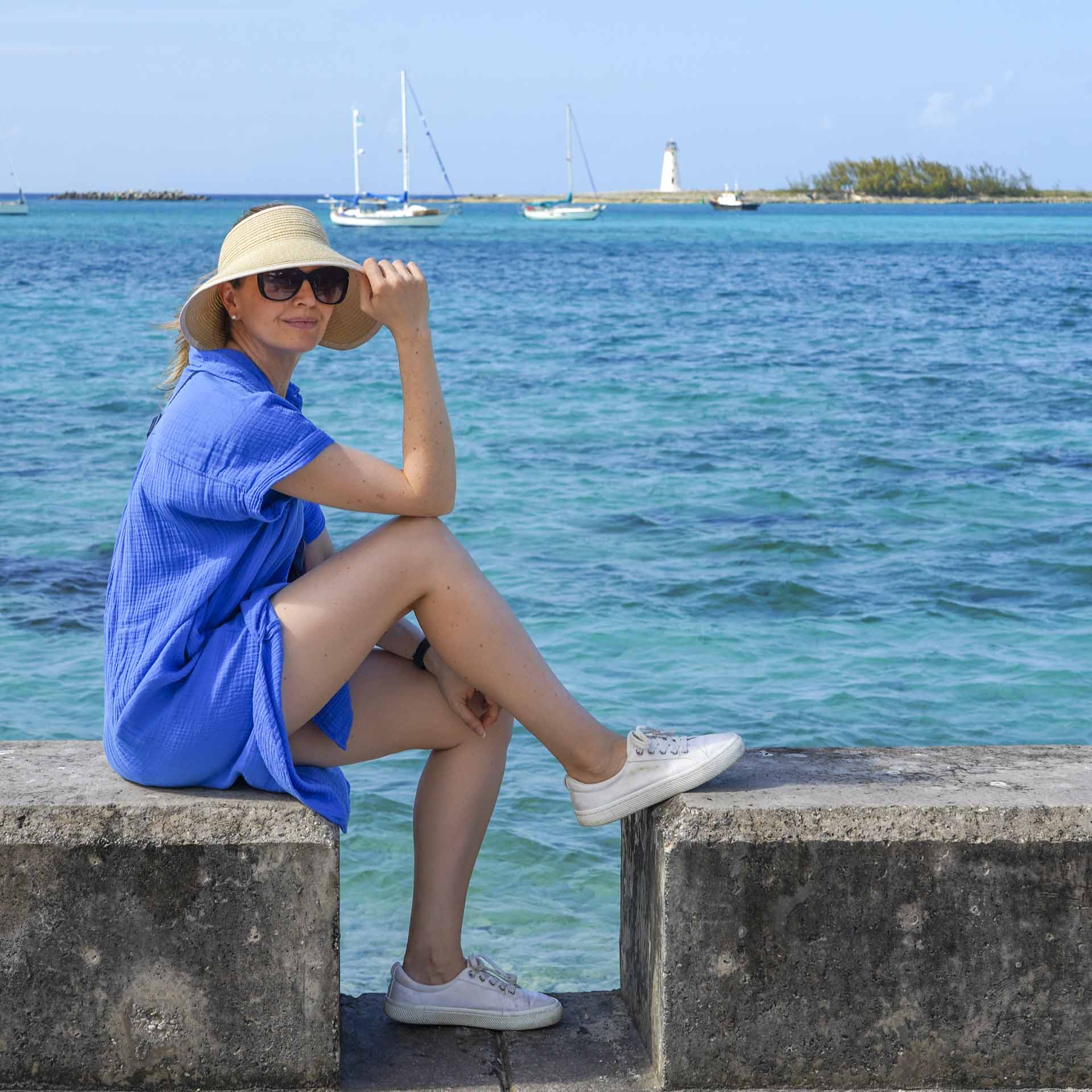 Madam ZoZo in blue dress and straw visor, seated on concrete block with Caribbean waters in the background. Moored yachts and a lighthouse are visible in the distance.